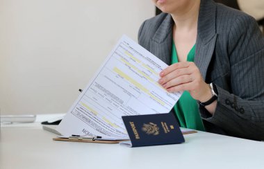 Kafkasyalı bir kadının elinde vize başvuru kâğıdı belgesi ve ofis masasında Amerikan pasaportu olan vize başvurusu konseptini yakın plan.