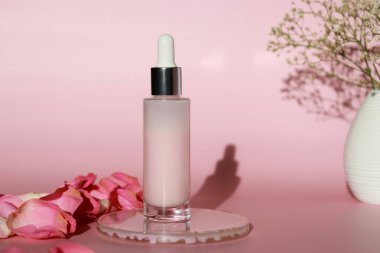 Pembe arka planda ve pembe yapraklarda gül özü olan bir şişe yüz serumu losyonu. Nemlendirici cilt bakımı ürün konseptini oluşturur