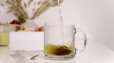 İçinde çay poşeti olan cam bardağa sıcak su dökmeye yakın. Arka planda rahat ev dekorasyonu olan beyaz masa 