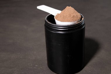 Bir kepçe dolusu çikolatalı protein tozunu siyah bir teneke kutuda, gri masa arkasında kapat. Besin takviyesi, beslenme 