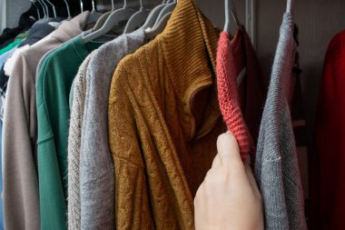 Dolaptaki askıların üzerinde kadın eli örgü süveter seçerken yakın plan. Rahat ve renkli kostüm konsepti