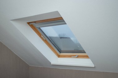 Beyaz ve bej duvarlı bir odada büyük bir Velux penceresinin içi, penceresinde gri gökyüzü 