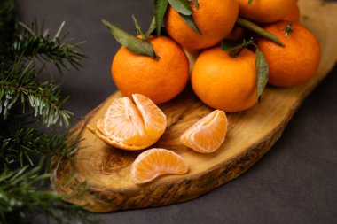 Soyulmuş mandalina dilimlerini ve ahşap tabakta bir yığın mandalinayı, önündeki bulanık yeşil çam dalını, gri masa arkasını kapatın.