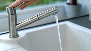 Mutfakta musluk açan bir kadının elini kapat. Beyaz lavabo ve paslanmaz çelik musluk. Arka planda yeşillikler olan bulanık bir pencere.