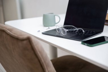 Şeffaf gözlükleri, dizüstü bilgisayarı, kahve fincanı ve malzemesi beyaz masada bulanık arka planı olan bir işyeri. Minimalist ve modern ofis ya da İçişleri Bakanlığı konsepti 