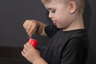 Kaşık ile yoğurt yiyen, gri gömlekli, arka planda bulanık gri duvar arkaplanı olan tatlı bir çocuğun yan portresi. 