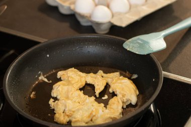 Çırpılmış omlet yumurtalarını siyah bir tavada mavi bir spatulayla kaplayın. Arka planda beyaz yumurtalar ve karton bir kutu.