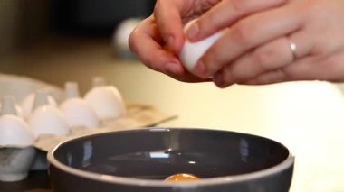 Kadın ellerinin beyaz bir yumurtayı bir kaseye kırarak omlet yumurtalarını tavada, bulanık mutfak masası arka planında beyaz yumurtalar yaparak kapatması. Yüksek kaliteli FullHD görüntüler