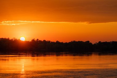 Nehir ya da göl üzerinde altın gün batımı. Güneş, ağaçların üstündeki ufka doğru batıyor. Resimli akşam manzarası