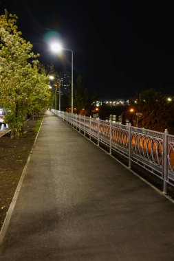 Asfalt yüzeyi ve güzel metal çitleri olan boş bir kaldırım. Yeni yaya yolu kutuplardaki sokak lambalarıyla aydınlanıyor.