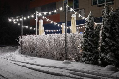 Gecenin geç saatlerinde kar yağdıktan sonra elektrik lambaları ve çitleri olan açık hava kafe alanı. Çalılarda kar var.