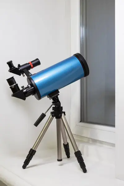 The telescope is on the windowsill and pointed out the window. Blue catadioptric astronomical telescope on a tripod in the room