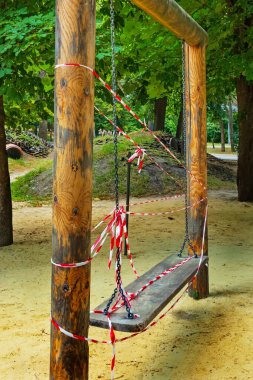 Wooden swing on chains on empty playground in park. White and red warning tape. Prohibited to use clipart