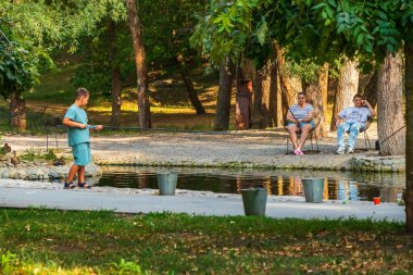 Kharkiv şehri, Ukrayna - 28 Ağustos 2024: Yetişkinler ve çocuklar bir şehir bahçesinde gölette balık tutuyorlar. Ellerinde olta, suyun yanında yerde demir kovalar. Bir yaz gününde şehirde amatör balıkçılık.