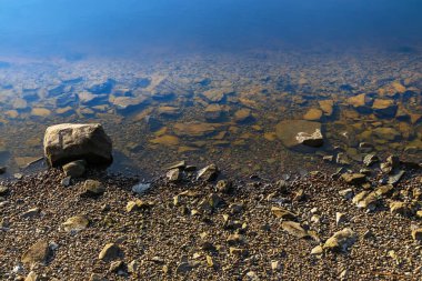 Suyun kenarında kayalık bir sahil var. Sahil şeridinde farklı boyutlarda sakin su ve taşlar var.
