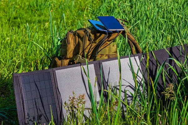 Taşınabilir güneş paneli sırt çantasının yanındaki yeşil çimlerin üzerinde katlanıyor. Akıllı telefonlar ve kişisel cihazlar için güneş enerjisi şarj istasyonu