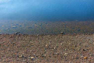Kıyıdaki küçük çakıl taşları durgun mavi suyun yanında
