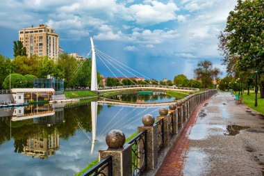 Harkiv nehri manzaralı şehir manzarası, yaya demir köprüsü ve binalar. Taştan çitlerle çevrili bir duvar. Bulutlu bir yaz günü, yağmurdan sonra
