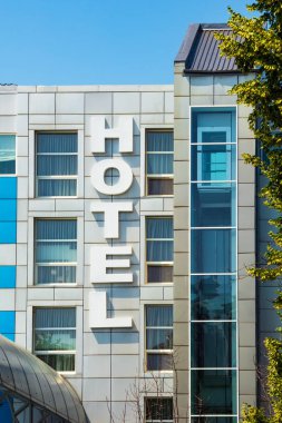 Büyük harfli oteli olan modern bir binanın dışı. Pencereleri ve asansörü olan çok katlı bir binanın ön cephesi.