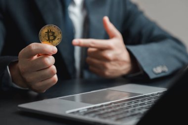 Takım elbiseli bir işadamı bilgisayarını işaret ederken elinde Bitcoin para tutuyor, modern yatırımlardaki teknoloji ve finans kavşağını gösteriyor..
