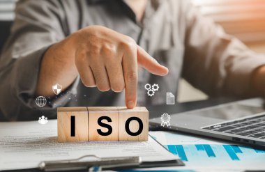 Bir işadamı, ofis masasında ahşap bloklar kullanarak, sertifika, kalite güvencesi ve etkili yönetim uygulamalarını temsil ederek ISO standartlarının seçimini gösterir..