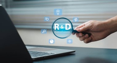 Bilgisayarın üzerinde büyüteç tutan bir el araştırma ve geliştirmeyi simgeler. Yenilik simgeleri, teknoloji içinde keşif ve analizi temsil eden objektifin etrafını sarar..