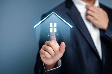 Takım elbiseli bir iş adamı, emlak ve ev sahipliğindeki modern teknolojiyi sembolize eden dijital bir ev ikonu ile etkileşime girer. Bu görüntü konut piyasasındaki yenilikleri ve dijital çözümleri temsil etmektedir..