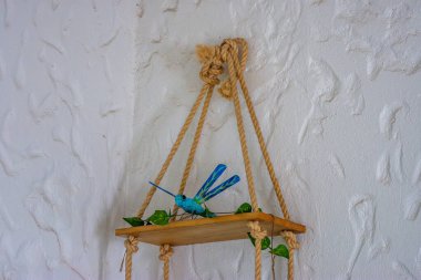 An artistic wooden shelf with a blue dragonfly decoration, surrounded by green leaves against a textured white wall