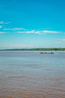 Amazon Nehri 'nde açık mavi bir gökyüzünün altında yemyeşil bir yeşil ile çevrili bir teknede iki balıkçıyı gösteren huzurlu bir manzara..