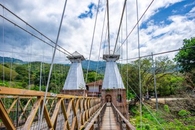 Santa F de Antioquia 'daki nehrin üzerindeki ikonik asma köprünün çarpıcı manzarası, yemyeşil bir zemin üzerinde koloni mimarisi sergiliyor..
