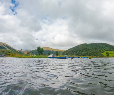 Laguna del Cumbal 'ın manzarası, bereketli dağlar ve sakin bir atmosfer ile çevrili, bulutlu gökyüzünün altında suda bir tekne sergileniyor.