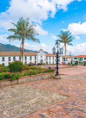 Mavi gökyüzü altında canlı çiçekler, palmiye ağaçları ve sömürge mimarisinin yer aldığı Mongui 'nin ana meydanının resimli görüntüsü.