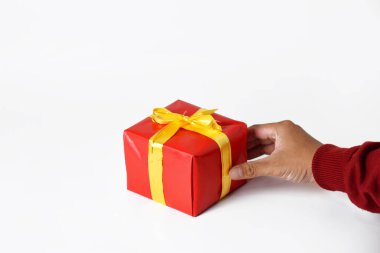 Kırmızı kazak giyen bir kişinin eli, beyaz arka planda sarı altın kurdeleli kırmızı bir hediye kutusu gösterir. Noel ve Yeni Yıl kutlamalarında