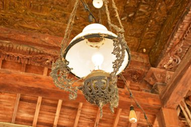 Beyaz cam gölgeli, antika, süslü bir lamba ve ahşap tavandan sarkan koyu renkli bir metal..