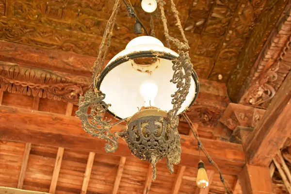 Beyaz cam gölgeli, antika, süslü bir lamba ve ahşap tavandan sarkan koyu renkli bir metal..