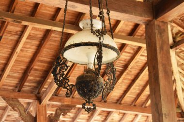 Beyaz cam gölgeli, antika, süslü bir lamba ve ahşap tavandan sarkan koyu renkli bir metal..