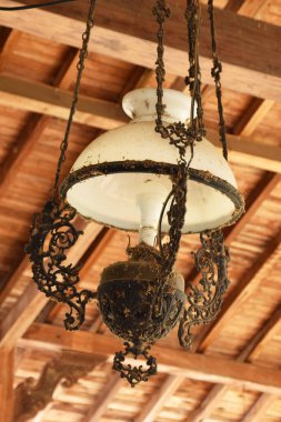 Beyaz cam gölgeli, antika, süslü bir lamba ve ahşap tavandan sarkan koyu renkli bir metal..