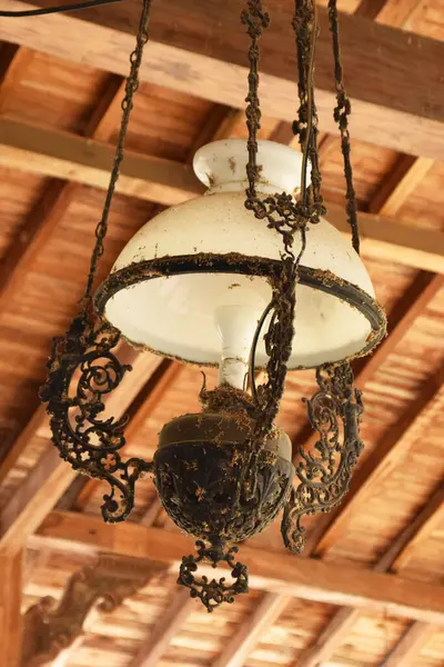 Beyaz cam gölgeli, antika, süslü bir lamba ve ahşap tavandan sarkan koyu renkli bir metal..