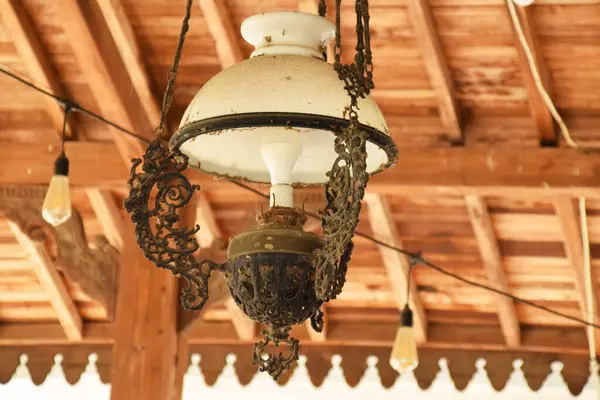 Beyaz cam gölgeli, antika, süslü bir lamba ve ahşap tavandan sarkan koyu renkli bir metal..