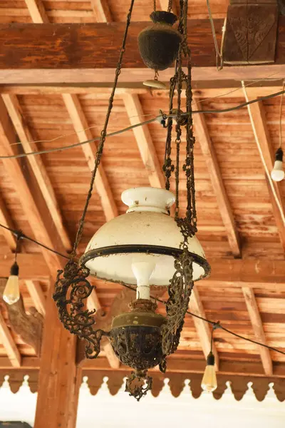 Beyaz cam gölgeli, antika, süslü bir lamba ve ahşap tavandan sarkan koyu renkli bir metal..