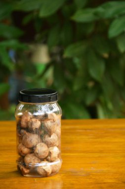 Siyah sarımsak karanfilleriyle dolu temiz cam bir kavanoz, yemyeşil yaprakların bulanık arka planıyla ahşap bir yüzeye oturur..