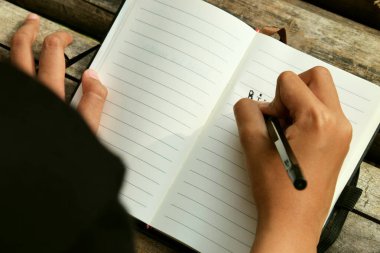 Bambu masasına yerleştirilmiş açık bir kitaba yazı yazan bir kızın üst görüntüsü. Doğa öğrenme, eğitim, yaratıcılık ve Dünya Kitap Günü temaları için mükemmel.