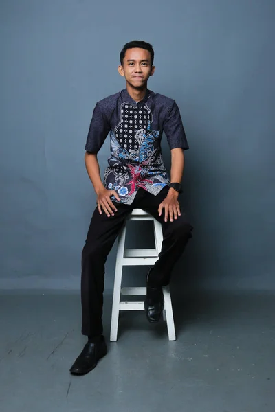 Successful and confident young businessman from asia sitting on chair. Asian man with dark hair wearing batik shirt with trousers and looking at the camera. Isolated on dark gray background. Studio shooting