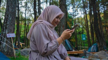 Açık havada akıllı telefon kullanan tesettürlü genç bir kadının fotoğrafı. Gülümseyen Endonezyalı kadın evde akıllı telefon kullanıyor, mesaj gönderiyor ya da parkta dinlenirken sosyal ağlara takılıyor