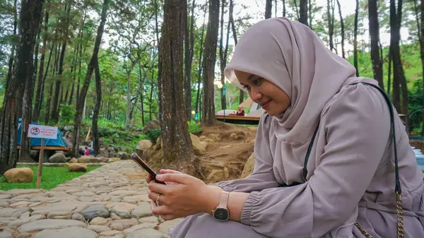 Açık havada akıllı telefon kullanan tesettürlü genç bir kadının fotoğrafı. Gülümseyen Endonezyalı kadın evde akıllı telefon kullanıyor, mesaj gönderiyor ya da parkta dinlenirken sosyal ağlara takılıyor