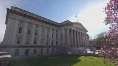 Washington, D.C. 'deki Hazine Bakanlığı' nın kuzey girişinde güneşli bir bahar günü görüldü. Ön planda çiçek açan pembe çiçekli bir ağaç görülüyor..