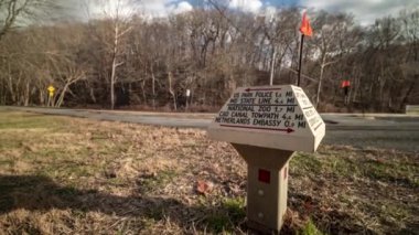 A time-lapse of a trail marker located near Beach Drive in Rock Creek Park in Washington DC. The camera rotates around the signpost, with motion-blurred people moving about in the background.