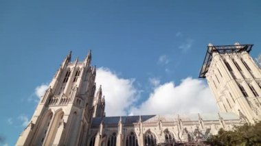 A time-lapse shot from a low angle pans across the south side of the Washington National Cathedral in Washington, DC. Clouds in the sky pass by the towers and scaffolding is seen on the facade.