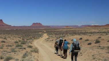 Bir grup gezgin Utah 'taki Glen Canyon Ulusal Dinlenme Bölgesi' nde toprak bir yolda yürüyüş yapıyor. Uzakta, Canyonlands Ulusal Parkı 'nda bulunan tasarruf ve kalçalar ve karlı zirveler görülüyor..
