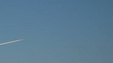 Bir jet uçağı mavi gökyüzünde çaprazlama uçarak turuncu güneş ışığı altında beyaz bir iz bırakır..